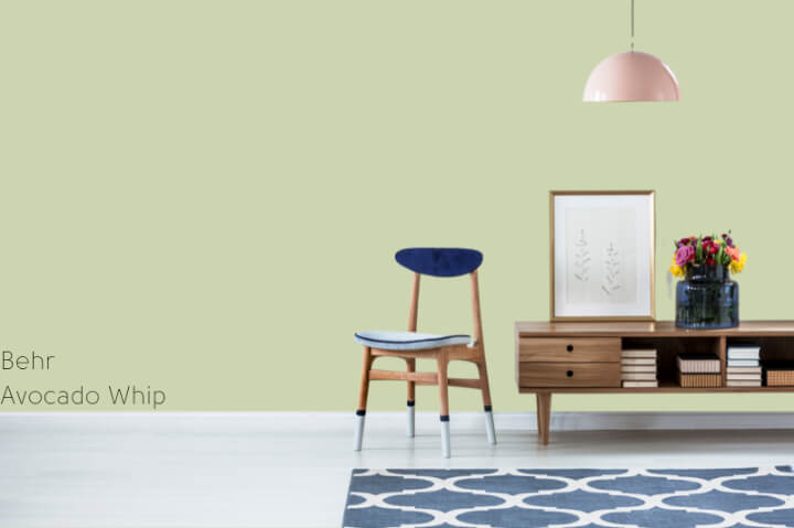 Avocado whip by Behr: a pale creamy green living room with a blue velvet and wood chair, a wood coffee table, blue rug, and a pink hanging light.