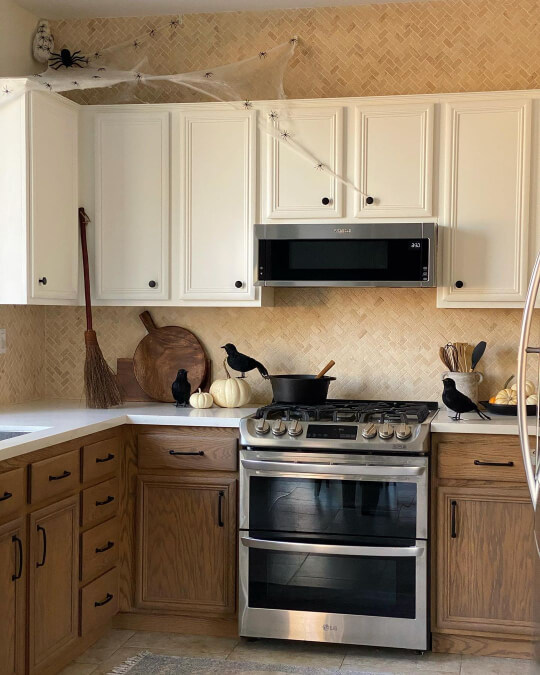 Creamy white upper cabinets with walnut mid-toned lowers, black hardware, white quartz countertops, and a beige mini herringbone tile backsplash. Kitchen is decorated for subtley for halloween with pumpkins fake crows and cobwebs.