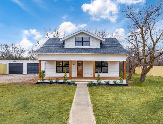 A look down the sidewalk towards a White exterior of a modern farmhouse with black roof, one large dormer, black windows, and natural wood trim and pillars