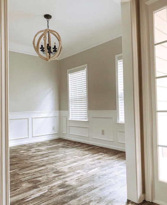 Paint of Behr Silver Drop on a room wall with white wainscoting on the lower half. Wooden chandelier display. Lights coming through the window blinds.