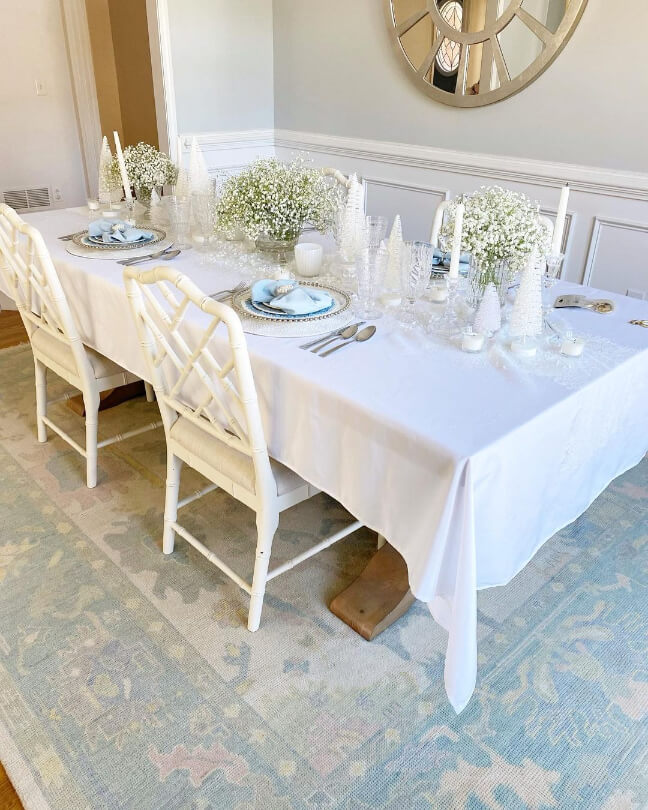 Light French Gray looks more gray on the upper portion of dining room walls with white wainscoting on the lower half. A large table with white tablecloth and white chairs sit on a coordinating tiffany blue patterned rug