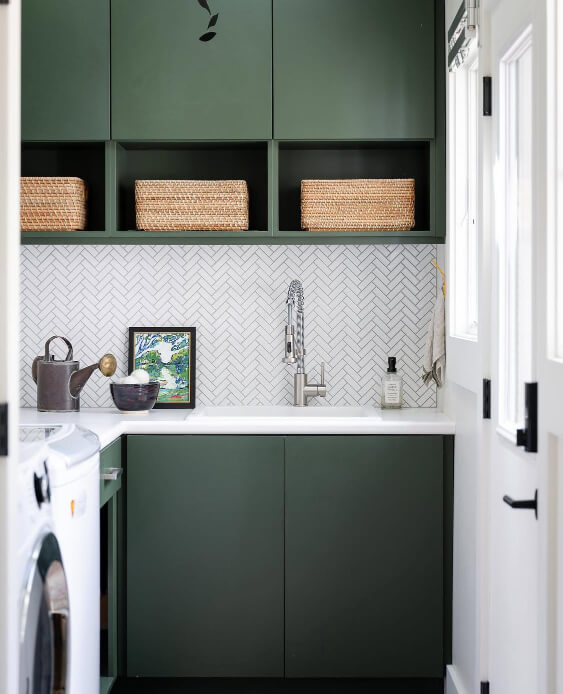 Backwoods cabinets are featured in a laundry room with a white countertop and a herringbone patterned tile backsplash above the sink.