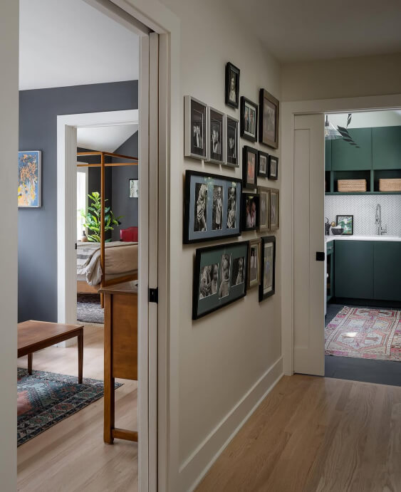Backwoods green cabinets and a herringbone patterned tile backsplash are visible in a laundry room through a doorway, off a hallway wall decorated with a gallery of framed photos.