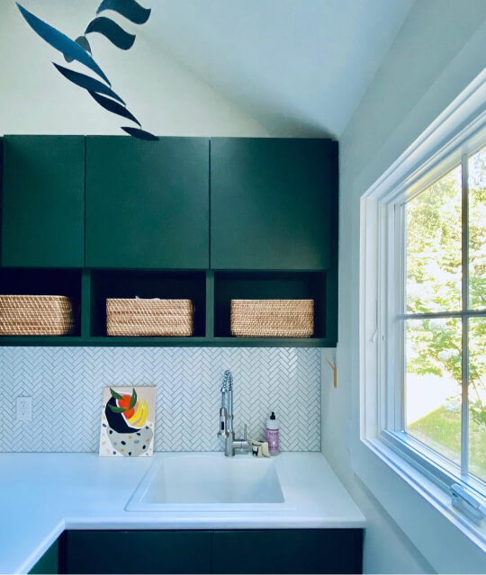 Backwoods green cabinets are mounted above a white sink, white countertop, and herringbone patterned tile backsplash in a bright laundry room with a large window and a vaulted ceiling.