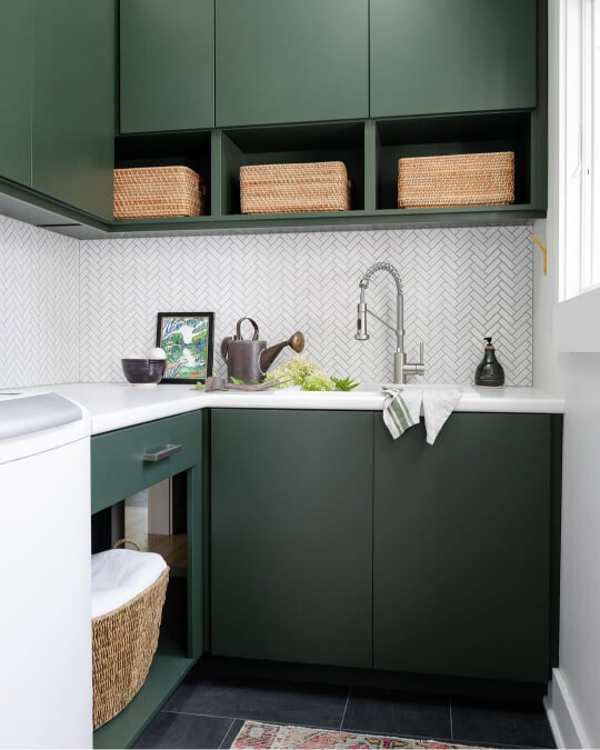 Backwoods green cabinets and upper open shelving are installed above a white countertop and a herringbone patterned tile backsplash in a modern laundry room.