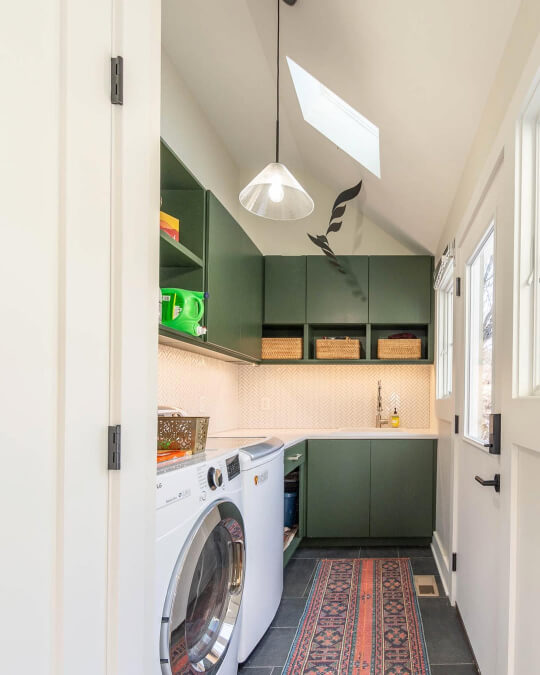 Backwoods cabinets and a herringbone patterned tile backsplash are installed above a washer and dryer in a long, narrow laundry room with a vaulted ceiling and a terracotta-colored persian patterned rug.