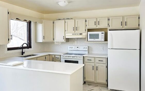 Benjamin Moore Revere Pewter Kitchen cabinets with recessed panel doors and black bar hardware, white solid-surface countertops, a white hexagon tile backsplash, a farmhouse-style sink under a window with a black faucet, white appliances including a stove, microwave, and refrigerator, and light marble-look tile flooring.