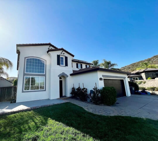 Creamy exterior paint covers a large, two-story Spanish-style house with dark trim and shutters, featuring a large arched window and a two-car garage with a dark panel patterned door under a sunny blue sky.