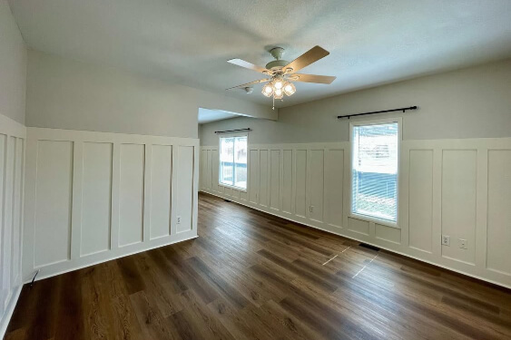 Creamy walls with white board and batten paneling cover a bright, empty room with dark wood-look laminate flooring, two windows, and a white ceiling fan with a chandelier light fixture.