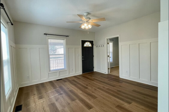 Creamy walls with white board and batten paneling cover a bright, empty room with dark wood-look laminate flooring, two windows, and a white ceiling fan with a chandelier light fixture.