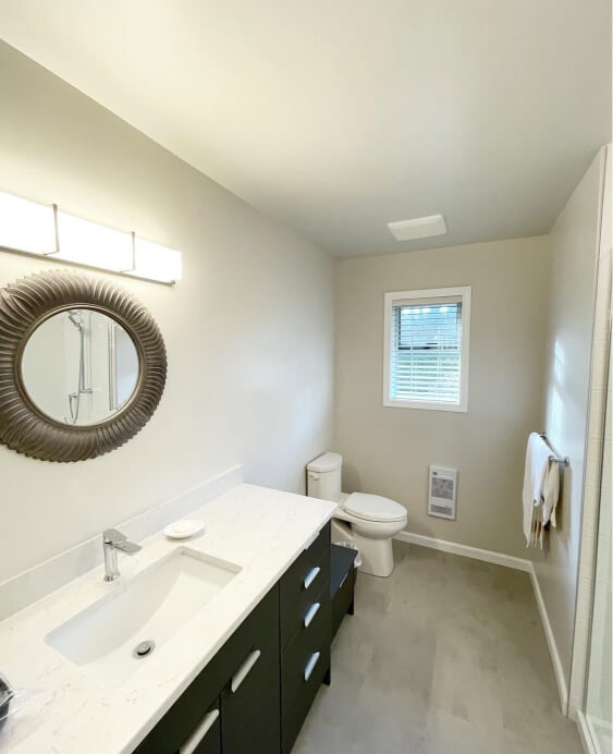 Pale Oak on the walls behind a modern bathroom vanity with a white countertop, a round decorative mirror, a toilet, a window, and a towel hanging on the right side.