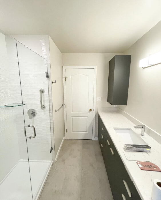 Pale Oak on the walls behind a glass shower enclosure, a white door, a long vanity with a white countertop, and a Matt Charcoal cabinet mounted above the counter.