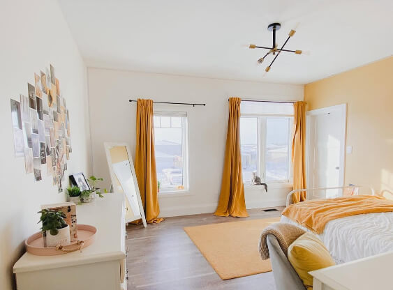 Pale Oak Wall and a Marblehead Gold accent wall. The room features two tall windows with golden curtains, a bed with yellow and white bedding, a white dresser with plants and decor, a standing mirror, and a modern black-and-gold ceiling light. Sunlight fills the space, creating a fresh and cozy atmosphere.