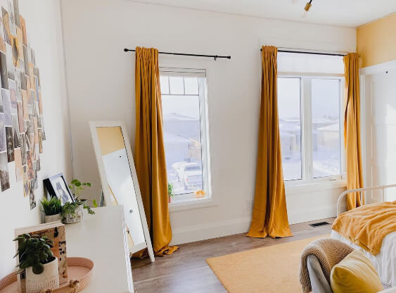 Pale Oak–painted wall, two tall windows with mustard-gold curtains, a leaning mirror beside a white dresser topped with plants and decor, and natural light filling the room. Snowy scenery is visible through the windows, creating a warm contrast with the cozy interior.