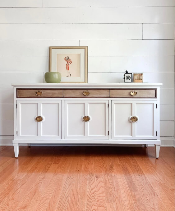 Pale oak cabinets on a white sideboard with brass knobs, decorated with framed art and small décor pieces in a bright room with wood flooring.