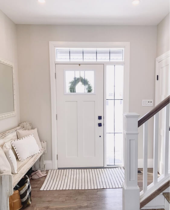 Pale oak walls surround a white front door in an entryway, which is accented by a striped rug and a white distressed bench with pillows.