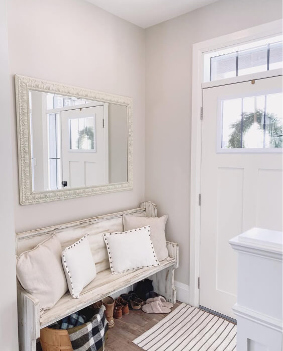 Pale oak walls serve as the backdrop for a distressed white bench with a white studded pillow and a matching ornate mirror in an entryway, where a white front door and a striped rug are also visible.