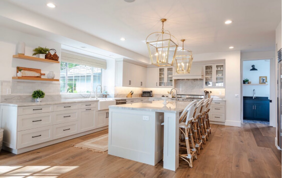 Pale Oak on cabinets surround a bright, spacious kitchen featuring white cabinetry, marble countertops, a large central island with wicker barstools, and three gold geometric pendant lights overhead. The room includes open wooden shelves, a farmhouse-style sink beneath a wide window, warm wood flooring, and a secondary prep area visible in the background.