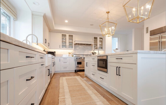 Pale Oak oon cabinets warms the walls of this bright, contemporary kitchen, complementing the white shaker cabinetry, marble countertops, and the long runner centered between the dual islands. Gold geometric pendant lights hang above, adding elegance, while stainless-steel appliances, glass-front upper cabinets, and natural wood floors complete the clean, airy look.