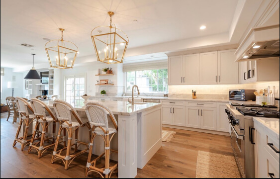 Pale oak cabinets in a bright, modern kitchen with a large marble island, rattan bar stools, gold pendant lights, and wide-plank wood flooring.