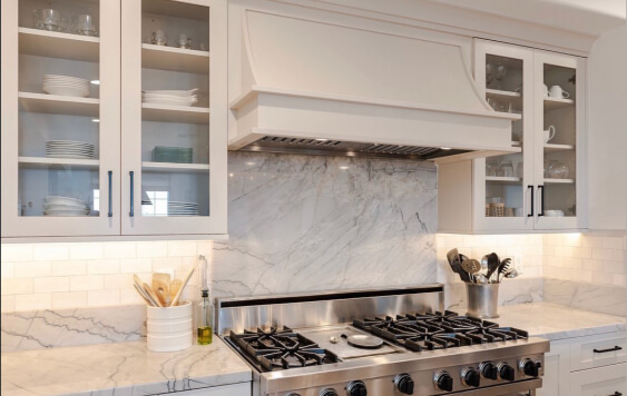 Pale oak cabinets around a stainless-steel stove with a marble backsplash, glass-front upper cupboards, and a bright, modern kitchen setup.